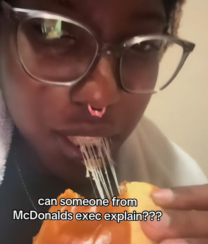 A person in glasses eating a hamburger with gooey cheese, questioning the McDonald's executive about fake foods Americans eat. A person in glasses eating a hamburger with gooey cheese, questioning the McDonald's executive about fake foods Americans eat.