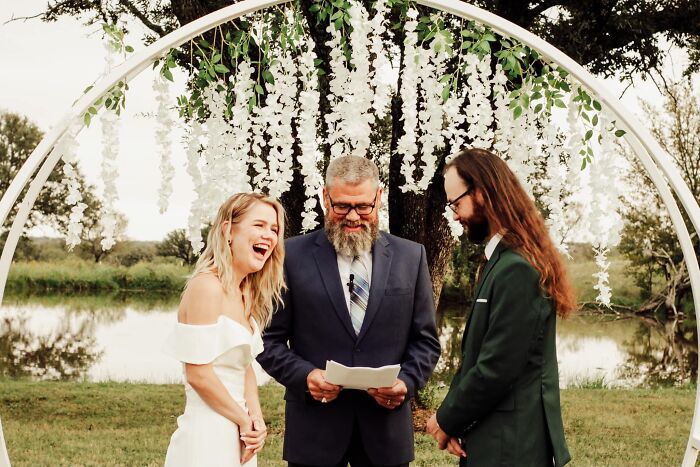 A bride laughs next to her groom and officiant under a floral arch in a funny wedding photo.