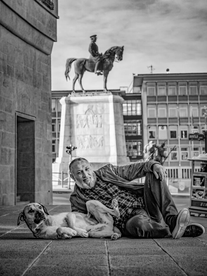 Mehmet Bayram street photo of a man and a dog lying on the pavement, with a historical statue in the background.