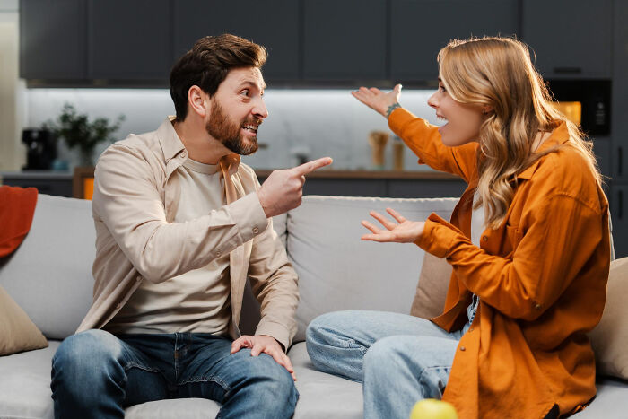 A man and a woman arguing intensely on a couch, pointing fingers, showing dumb reasons couples divorce.