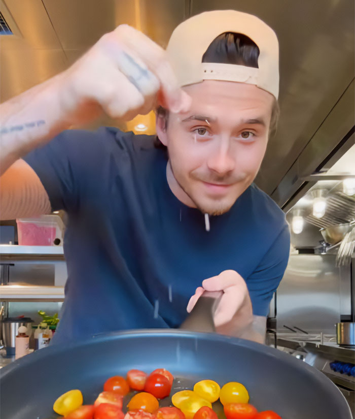 Brooklyn Beckham preparing a cooking video, adding ingredients to a pan with cherry tomatoes.