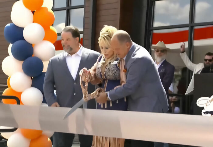 Dolly Parton and two men cutting a ribbon at her last public appearance, amidst conspiracy theory buzz. Dolly Parton and two men cutting a ribbon at her last public appearance, amidst conspiracy theory buzz.