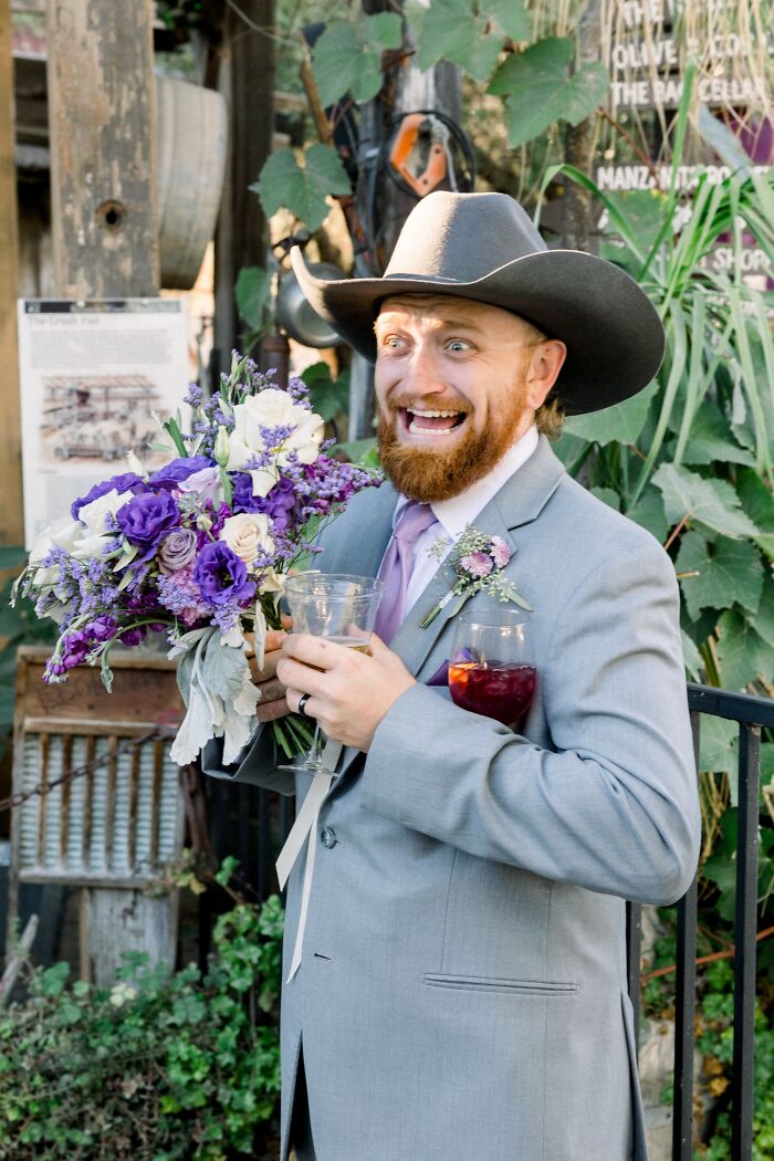 A groom in a cowboy hat holding flowers and drinks, making a funny wedding photo face.
