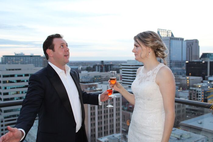 An iconic funny wedding photo of a bride and groom toasting with champagne on a rooftop overlooking a city.