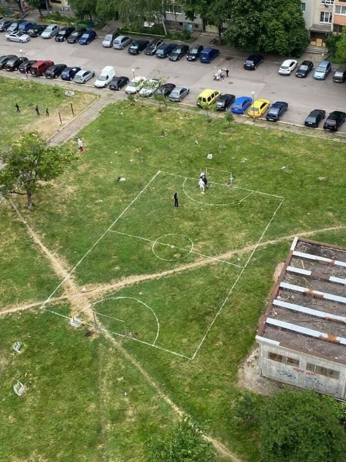 An aerial view of a makeshift football field in Eastern Europe, with lines drawn on a grassy area, surrounded by parked cars and buildings.