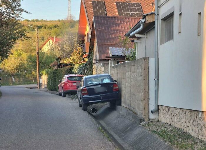 A blue car with its back wheel in a ditch next to a wall on a street in Eastern Europe, showing why people are built different.