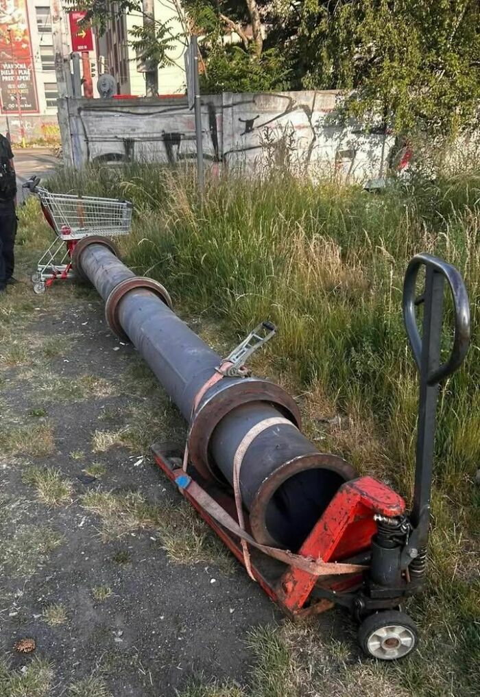 A very long pipe being transported on a hand pallet truck and a shopping cart in Eastern Europe, demonstrating improvised transport.