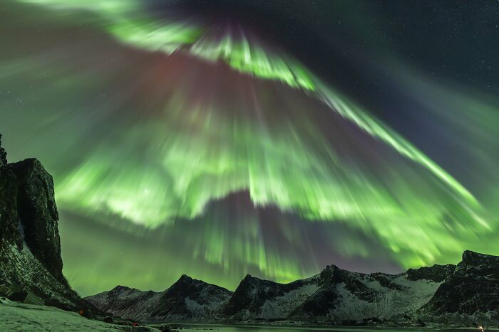 Breathtaking astronomy photo of brilliant green aurora rays illuminating the night sky over snowy peaks and water.