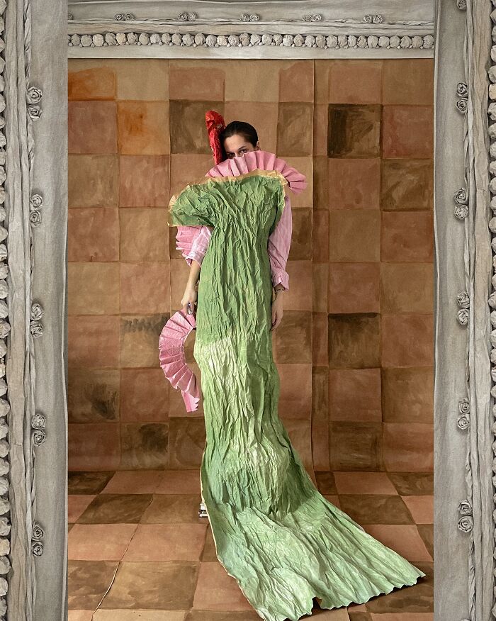 A person in a flowing green paper creation with pink ruffles stands in a decorative frame, blurring fashion and sculpture.