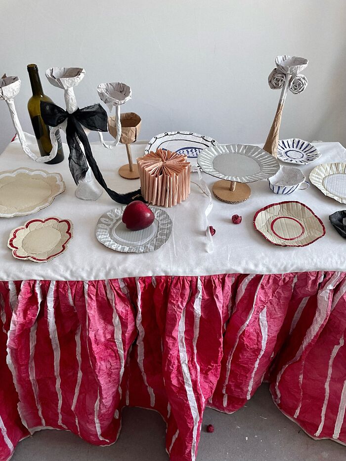Elaborate table setting with many surreal paper creations, including plates, candelabras, and a cake, resembling fashion.