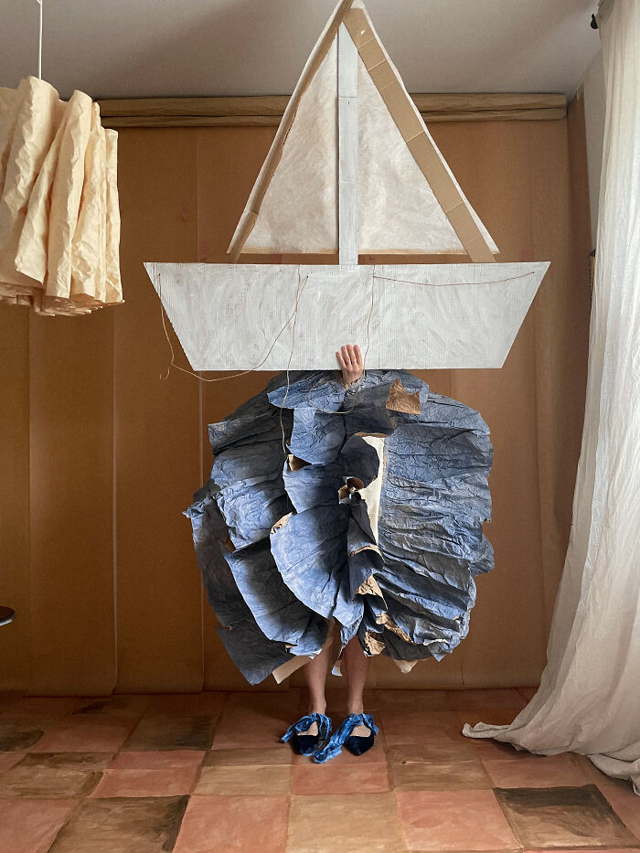 Artist in a paper boat creation and blue crumpled paper skirt, demonstrating surreal paper creations as sculpture.
