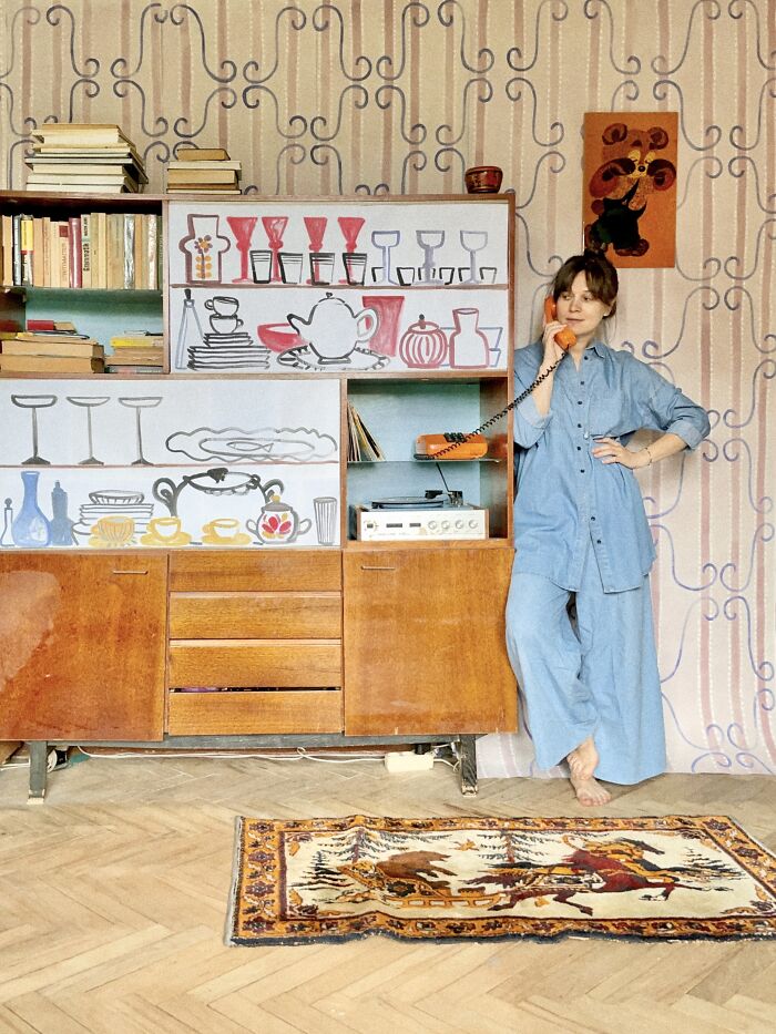 A woman in a blue paper outfit stands by a vintage cabinet, embodying surreal paper fashion and photography.