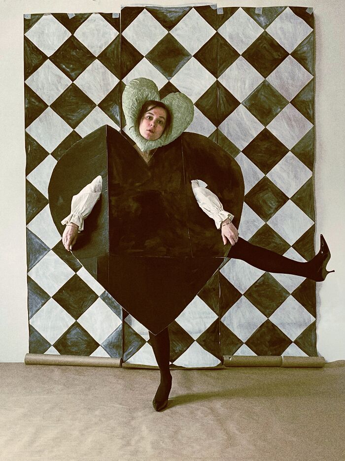 Artist in a black paper heart creation against a checkerboard background, showcasing surreal paper creations and fashion.