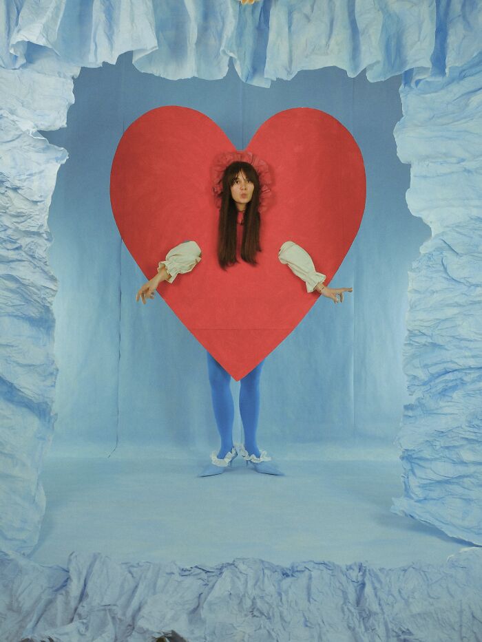 Surreal paper creation: a model inside a large red heart, framed by blue ruffled paper, showcasing fashion and sculpture.