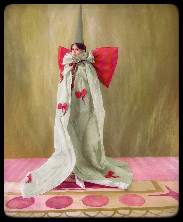 Surreal paper creation of a model wearing a long, light green paper gown with red bows and a tall hat, blending fashion and sculpture.