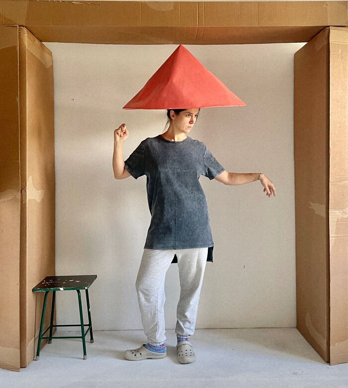 Artist wearing a large, red conical paper creation as a hat, blurring lines between fashion, sculpture, and photography.