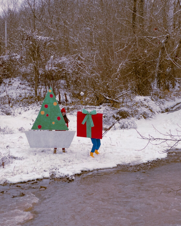 Two figures in surreal paper creations resembling a Christmas tree and a gift box in a snowy landscape, blurring fashion and sculpture.