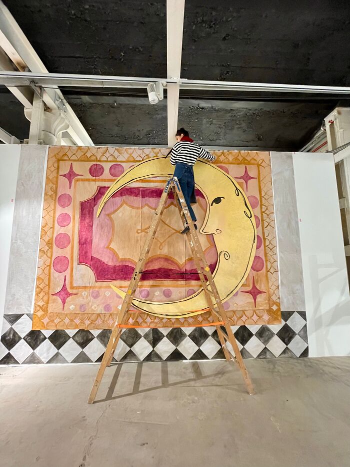 An artist on a wooden ladder applies finishing touches to a large paper creation featuring a crescent moon design, blurring sculpture and photography.