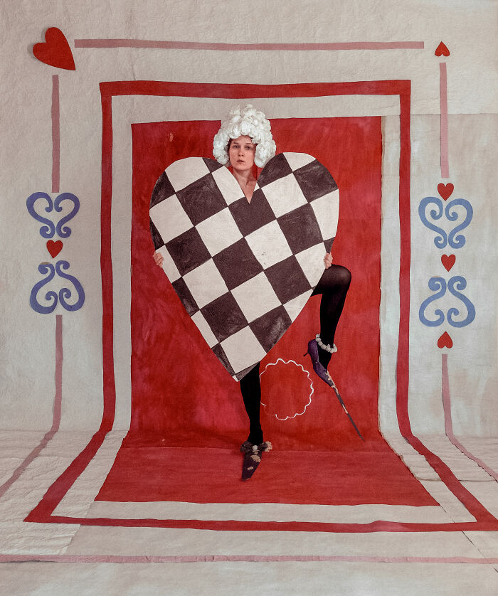 A person with a white wig and black tights holds a large, checkered heart, showcasing surreal paper creations and fashion.