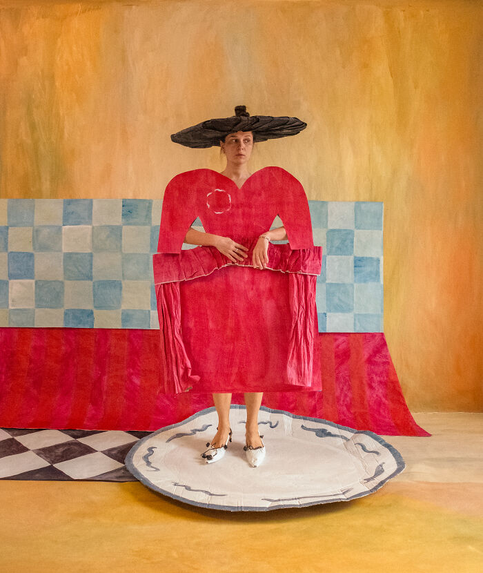 A person in a striking red heart-shaped paper creation with a black hat, standing on a white plate, showcasing paper fashion.