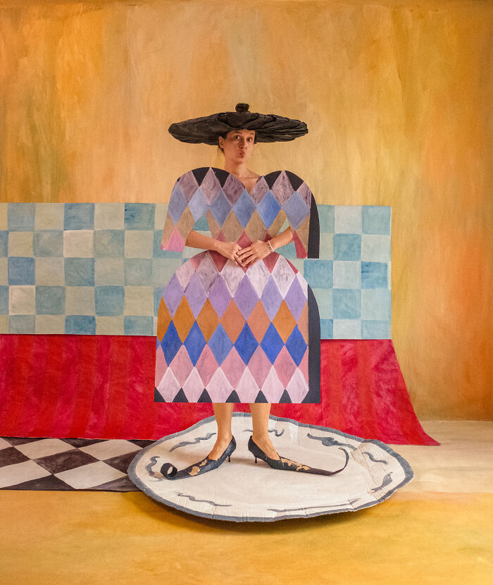 A person in a colorful harlequin-patterned paper creation with a large black hat, standing on a white plate, showcasing paper fashion.