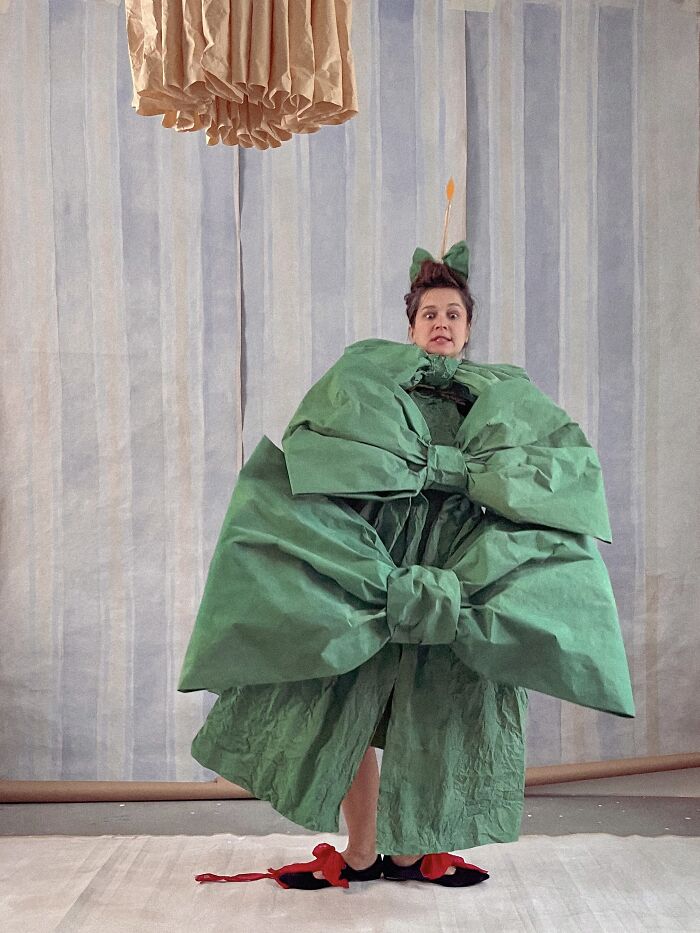 A person in a whimsical green paper bow creation stands against a light striped background, showcasing paper fashion and sculpture.
