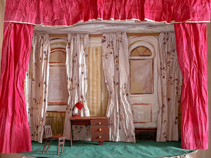 A detailed miniature room with red curtains, a desk, and a chair made from paper creations, blurring fashion and sculpture.