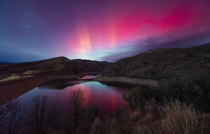 Stunning astronomy photo showcasing a bright pink and red aurora reflecting over a tranquil lake with hills in the background.