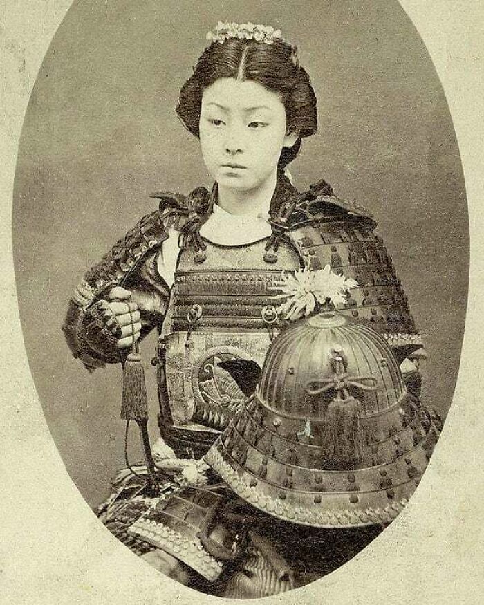 A vintage photo of a young woman in samurai armor with a helmet beside her, a moment from history.