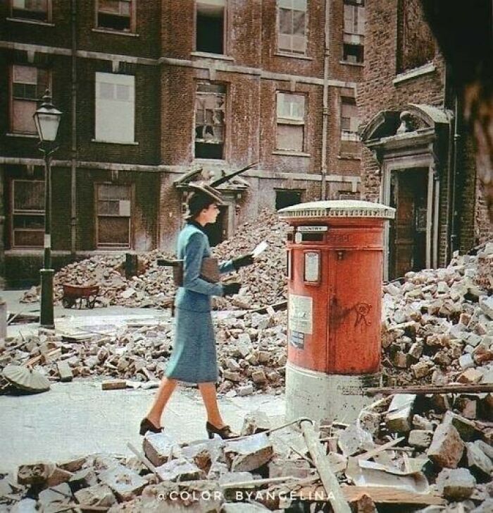 A woman in a stylish blue suit walks past a red post box amidst a landscape of rubble and destroyed buildings, a moment nearly lost to history.