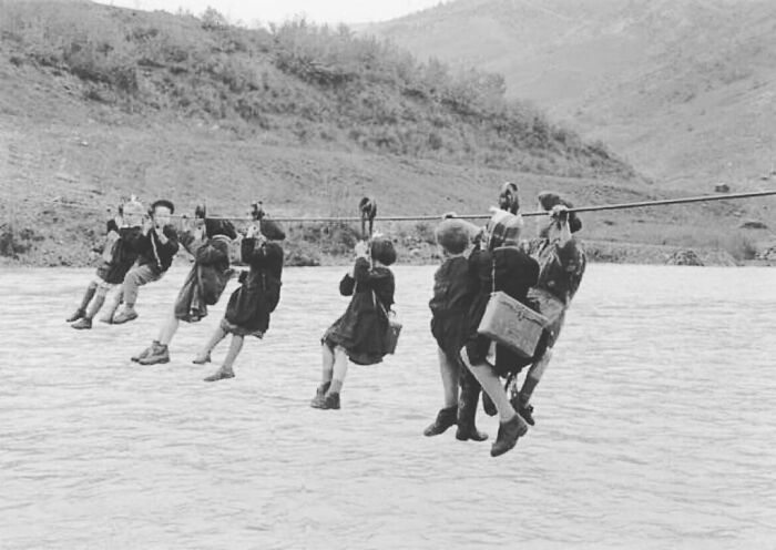 Black and white photo of children using a zipline to cross a wide river, a unique moment nearly lost to history.