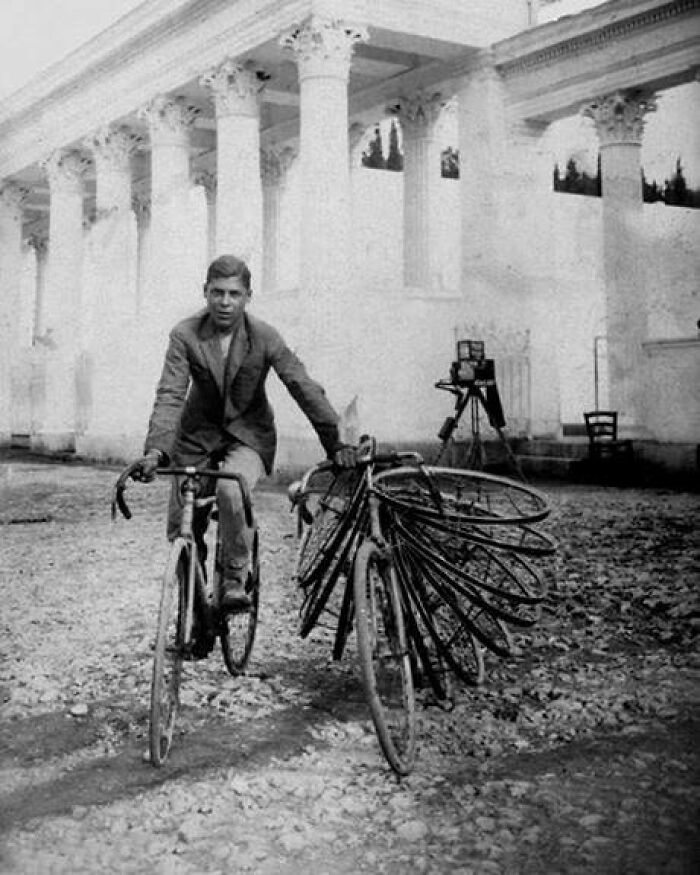 A man rides a bicycle with several spare wheels, in front of a classical building; a moment lost to history.