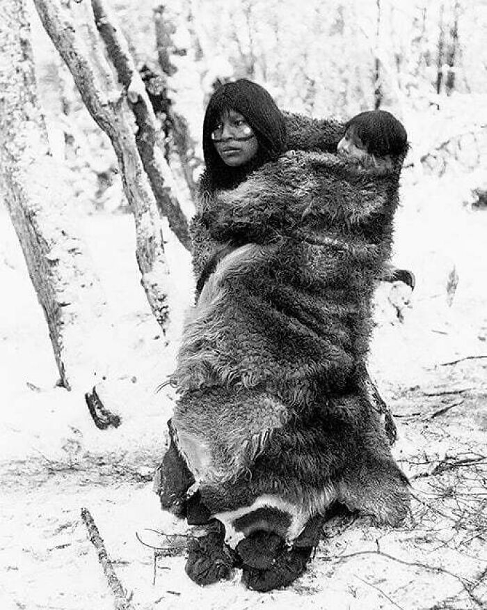 An indigenous person wrapped in furs carrying a child through a snowy forest, a cultural moment lost to history.