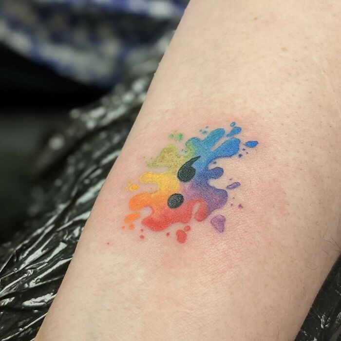 A vibrant rainbow watercolor splash semicolon tattoo on skin, a small symbol with a powerful story.