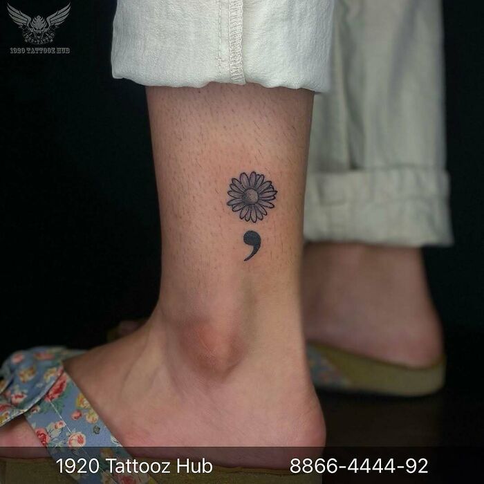 A simple black daisy and semicolon tattoo on an ankle, representing a small symbol with a powerful story.