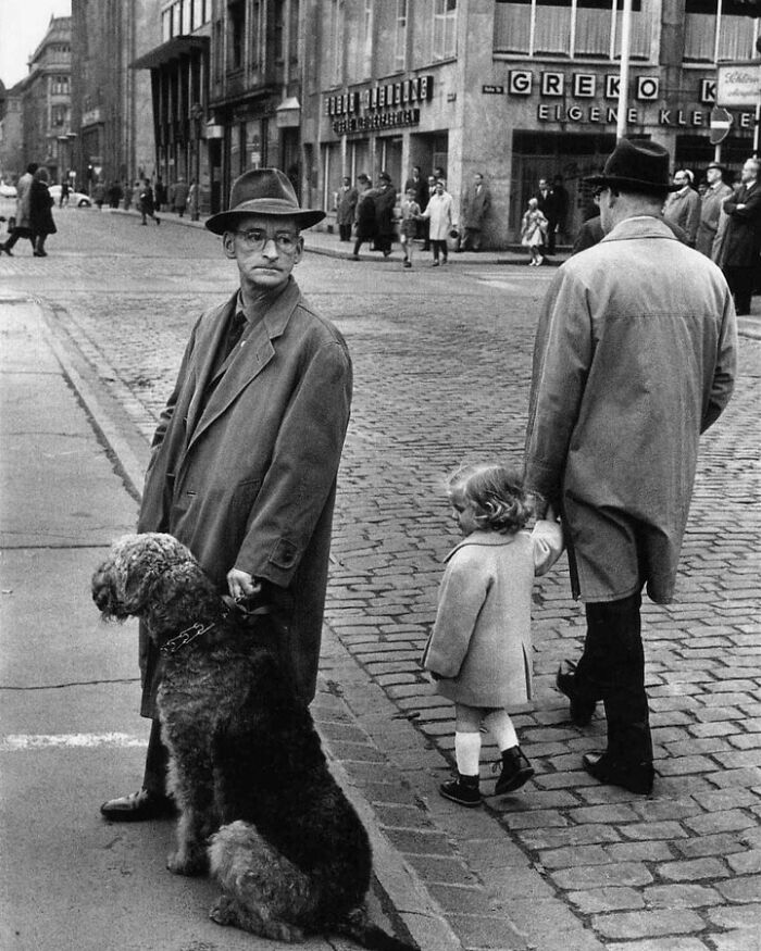 A man with glasses and a hat stands with a large dog while a child walks with another man on a cobblestone street, a moment nearly lost to history.
