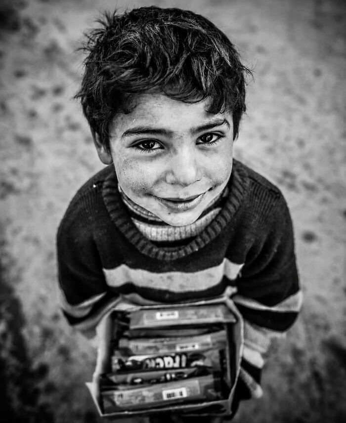 A black and white perfectly timed street photo by Mehmet Bayram shows a young boy holding a box of candies, smiling at the camera.
