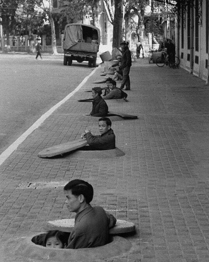 A line of people, including adults and a child, are partially submerged in sidewalk holes, observing a street scene with a truck, a moment nearly lost to history.