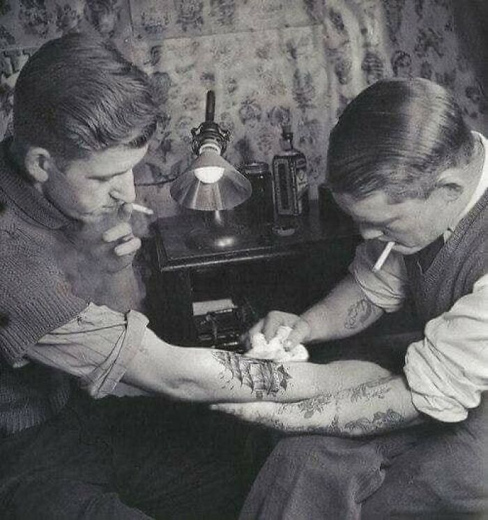 Two men in a vintage setting, one getting a tattoo, a candid moment nearly lost to history of early tattooing practices.