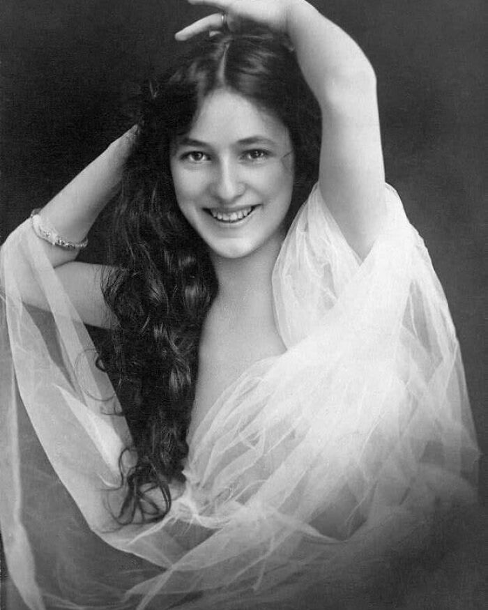 A black and white portrait of a young woman with long, dark hair, smiling and draped in sheer fabric, a moment nearly lost to history.