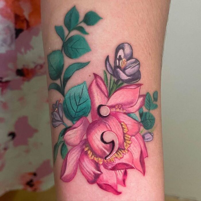 A colorful semicolon tattoo of pink flowers and green leaves on an arm, representing a small symbol with a powerful story.