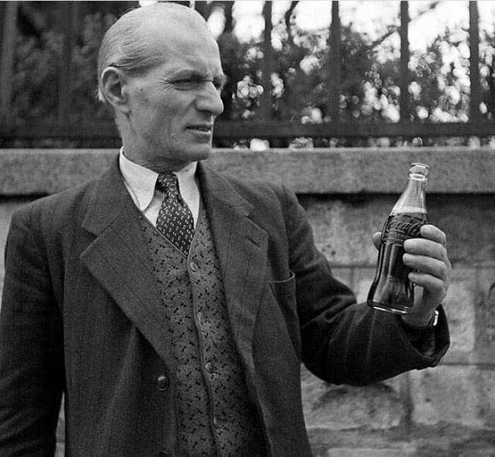 A man in a suit and tie holds up a bottle of Coca-Cola, looking to the side, capturing a moment nearly lost to history.