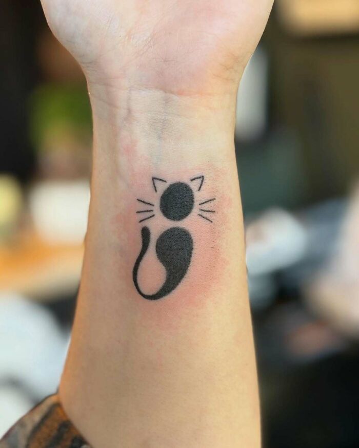 A minimalist black cat semicolon tattoo on a wrist, a small symbol with a powerful story.