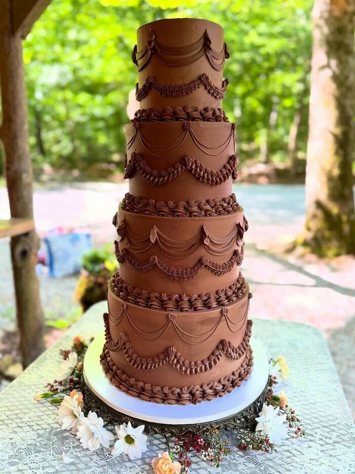 A tall, multi-tiered chocolate wedding cake decorated with elegant swags and piping, set outdoors.