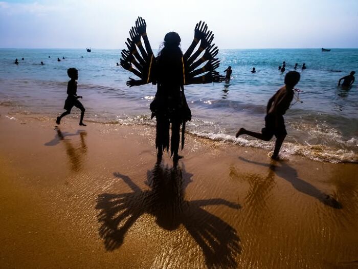 Children running on a beach past a person in an elaborate costume, a photograph by Tittu Shaji Thomas capturing life.