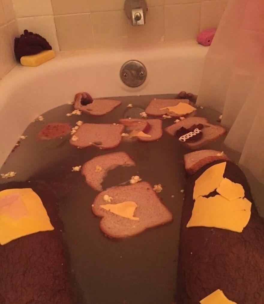 A person soaking in a bathtub filled with dirty water, pieces of bread, cheese, and popcorn, creating a cursed image.