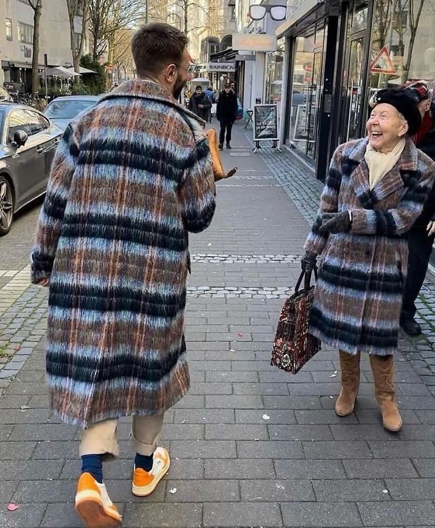 A man and an elderly woman wearing matching cursed plaid coats, laughing on a sidewalk, making eye contact.