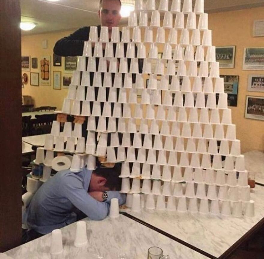 An unhinged, cursed image of a man building a tower of plastic cups around another sleeping man.