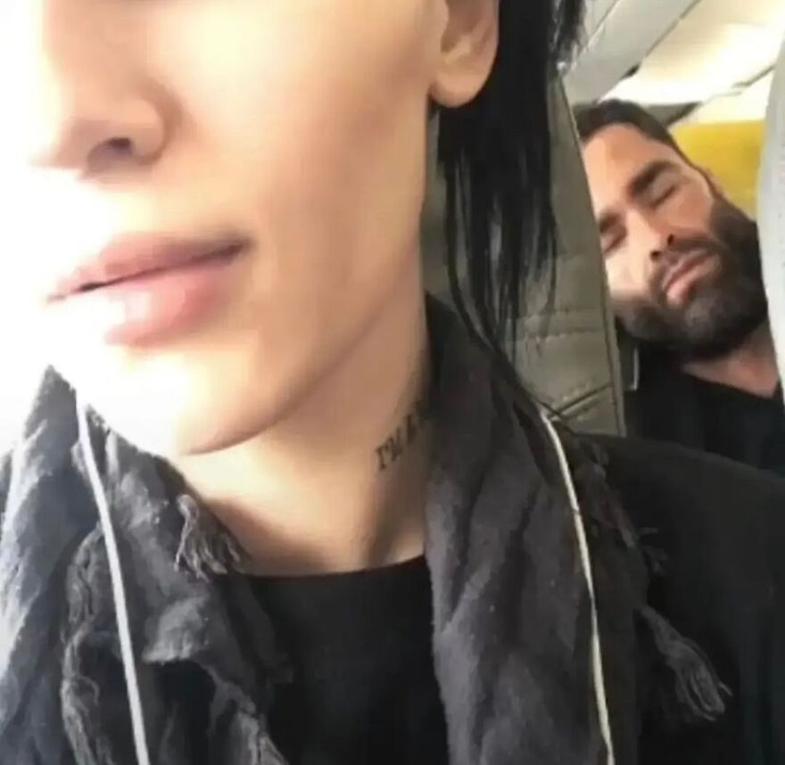 A cursed, unhinged image of a man sleeping on a plane behind another person taking a selfie.