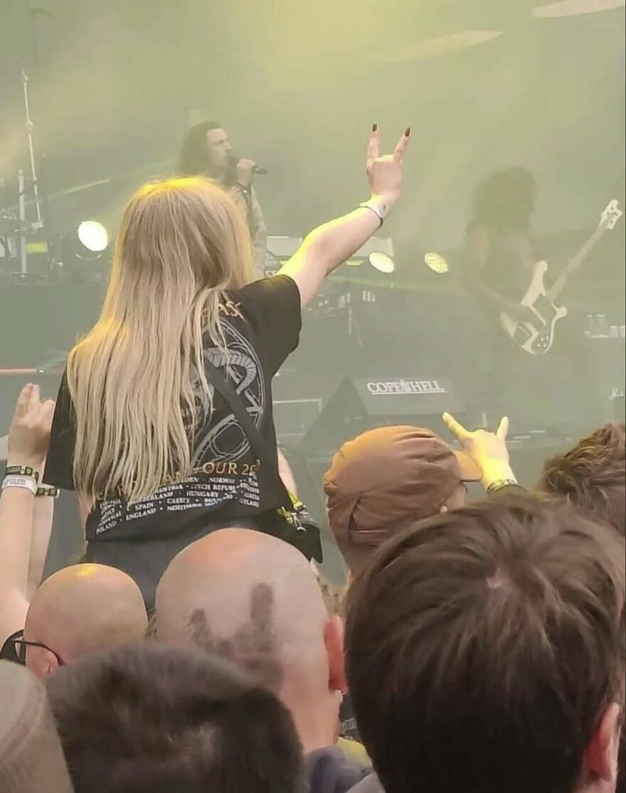A cursed image of a woman at a concert, making a rock hand gesture, with random lights and crowd in front of stage.